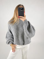 En person iført en grå sweater tager en selfie med en sort smartphone. Personen har hvidt tøj på og rød neglelak. Baggrunden er ensfarvet hvid.