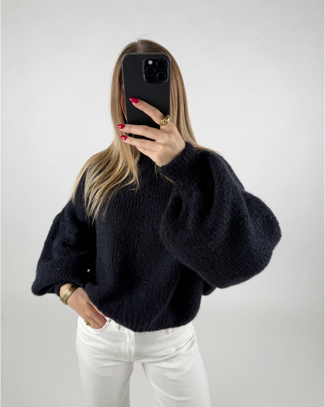En person iført en sort sweater holder en mobiltelefon op foran ansigtet og tager et selfie. Personen står i et rum med hvid baggrund, og har hvide bukser på.