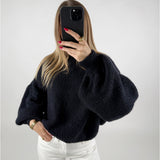 En person iført en sort sweater holder en mobiltelefon op foran ansigtet og tager et selfie. Personen står i et rum med hvid baggrund, og har hvide bukser på.