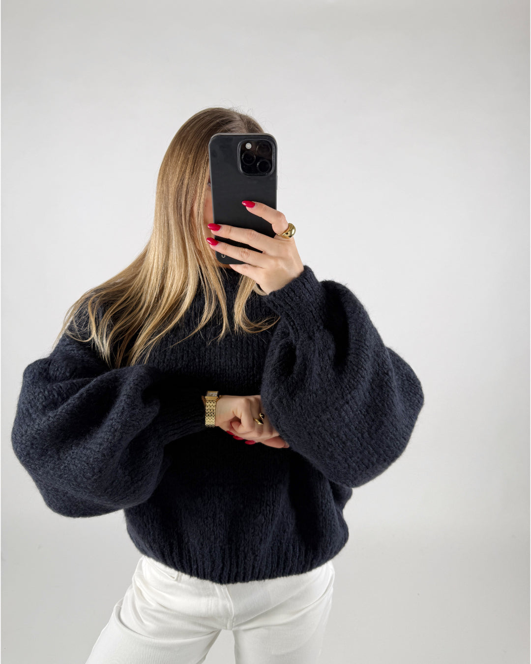 En person i en mørk sweater holder en smartphone op foran ansigtet. Personen har langt, lyst hår og bærer hvidt tøj og tilbehør i guld. Baggrunden er ensfarvet.