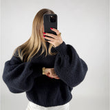 En person i en mørk sweater holder en smartphone op foran ansigtet. Personen har langt, lyst hår og bærer hvidt tøj og tilbehør i guld. Baggrunden er ensfarvet.