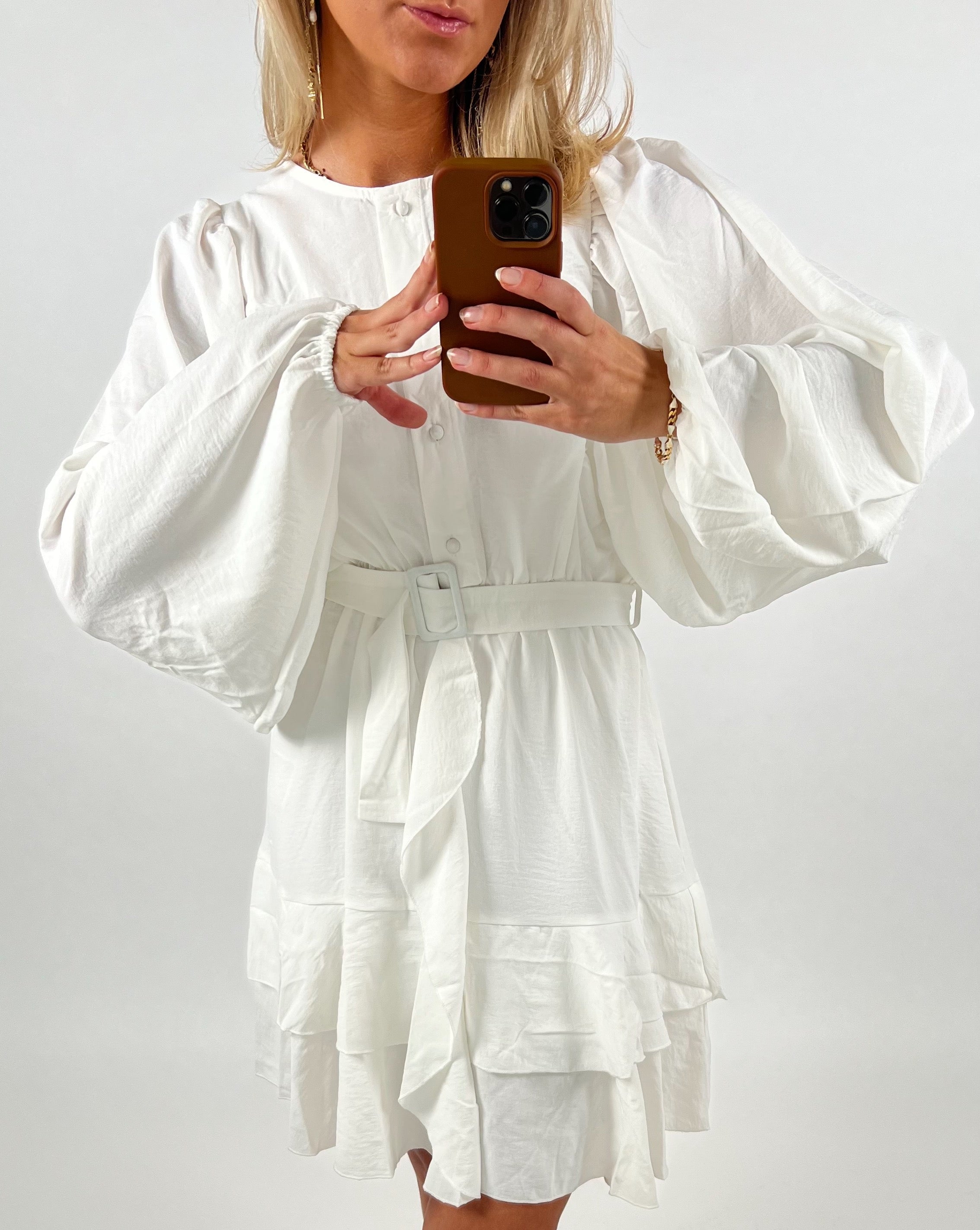 Person iført hvid kjole med brede ærmer og bælte taler et selfie. Kjolen har knapper, og personen står i et lys rum med en telefon i hånden.