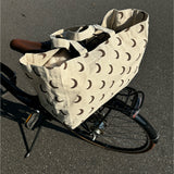 En beige stofpose med brune halvmåner, placeret på bagagebæreren af en cykel, står parkeret på en asfaltvej.