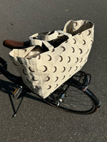 En beige stofpose med brune halvmåner, placeret på bagagebæreren af en cykel, står parkeret på en asfaltvej.