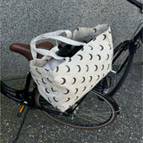 En stofpose med halvmånemønster hviler på en cykelbagagebærer. Cyklen står parkeret på en grusbelagt overflade op ad en stenvæg.