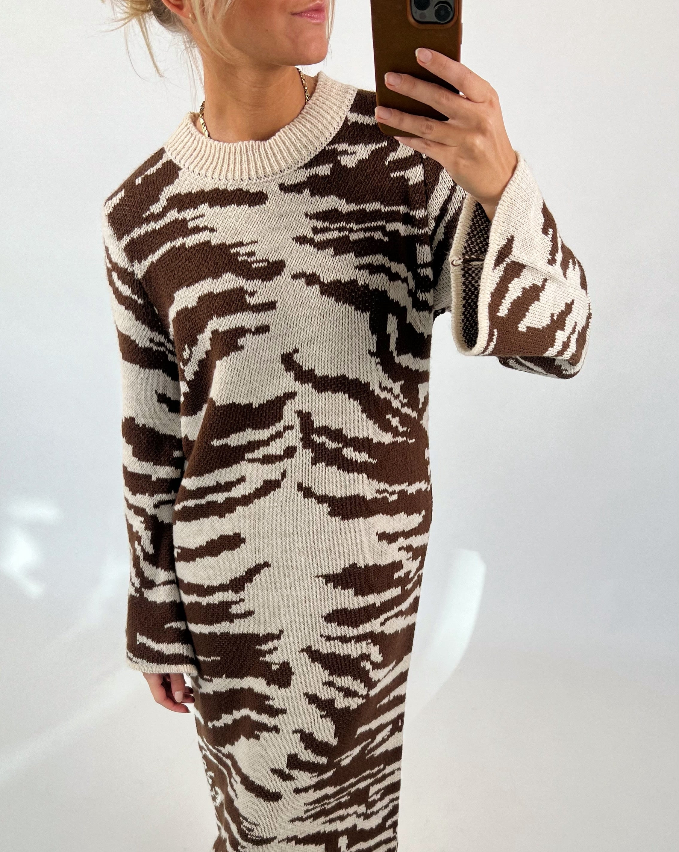 Kvinde i brunt og beige mønstret kjole tager en selfie med en smartphone i en minimalistisk, hvid baggrund.
