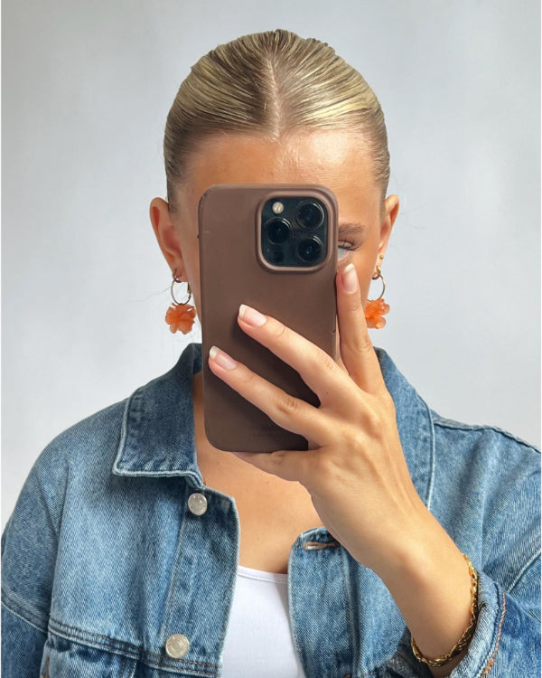 En person i en denimjakke holder en smartphone op foran ansigtet og tager et selfie. De har håret stramt redt tilbage og bærer øreringe med orange blomsterformede vedhæng.