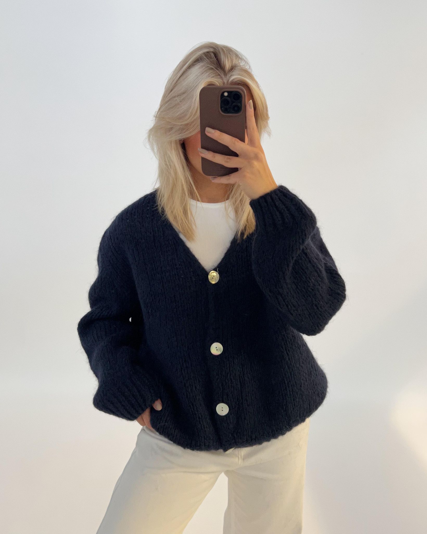 Person iført mørk cardigan med store knapper og lyse bukser, tager et spejlbillede i et neutralt oplyst rum. Personen dækker ansigtet med en smartphone.