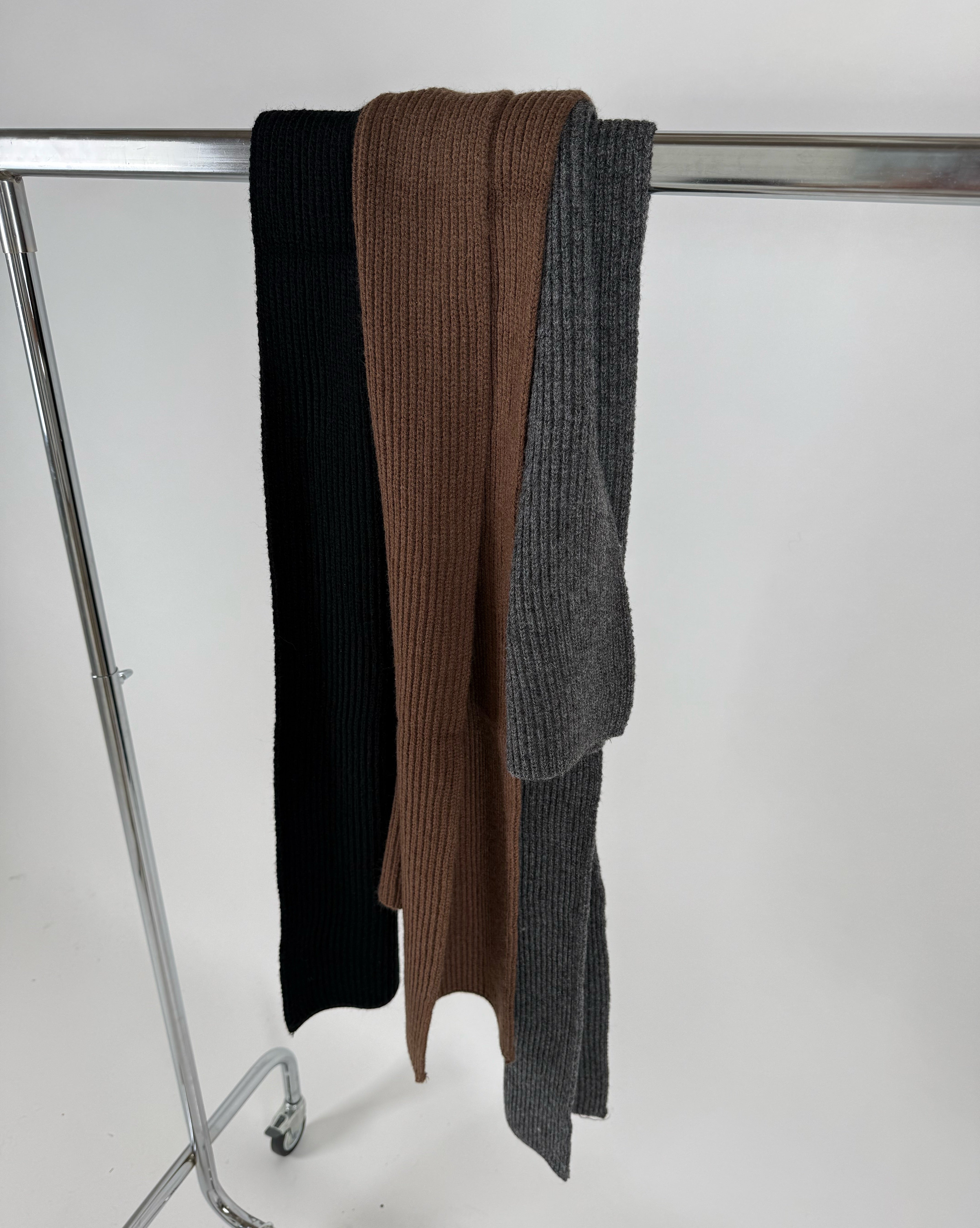 Tre ribstrikkede tørklæder, i farverne sort, brun og grå, hænger fra en sølvfarvet stang i en minimalistisk, lys baggrund.