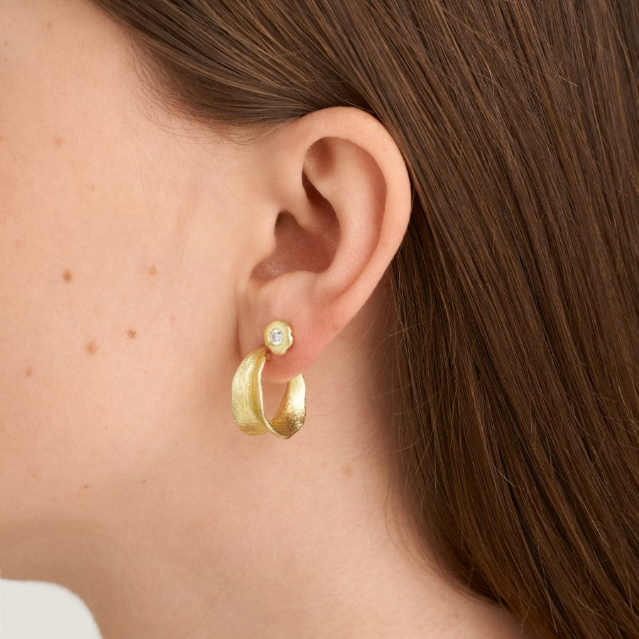 En gylden ørering med et lille indfattet ædelsten bæres på et øre, omgivet af brunt hår. Øreringen består af en elegant, buet form med en blank finish.