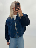 En person iført en denimjakke og lys denimnederdel tager et spejlbillede med en smartphone i et neutralt rum.