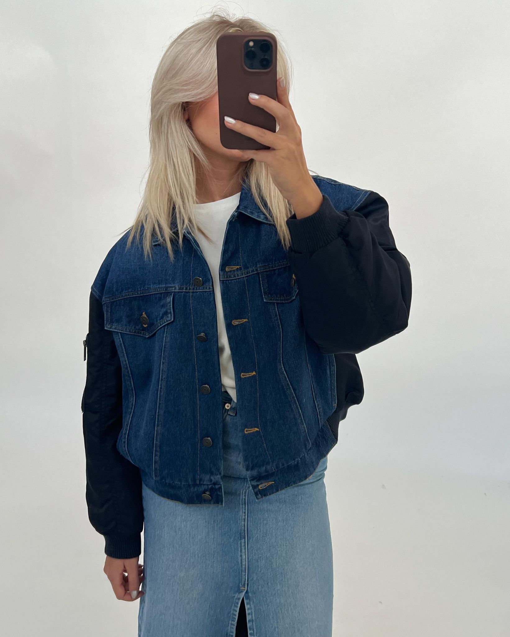 En person iført en denimjakke og lys denimnederdel tager et spejlbillede med en smartphone i et neutralt rum.