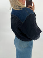 En person står iført en sort og blå jakke og lyst hår, tager et spejlbillede med en smartphone. Personen bærer også en lyseblå denim nederdel. Baggrunden er hvid.