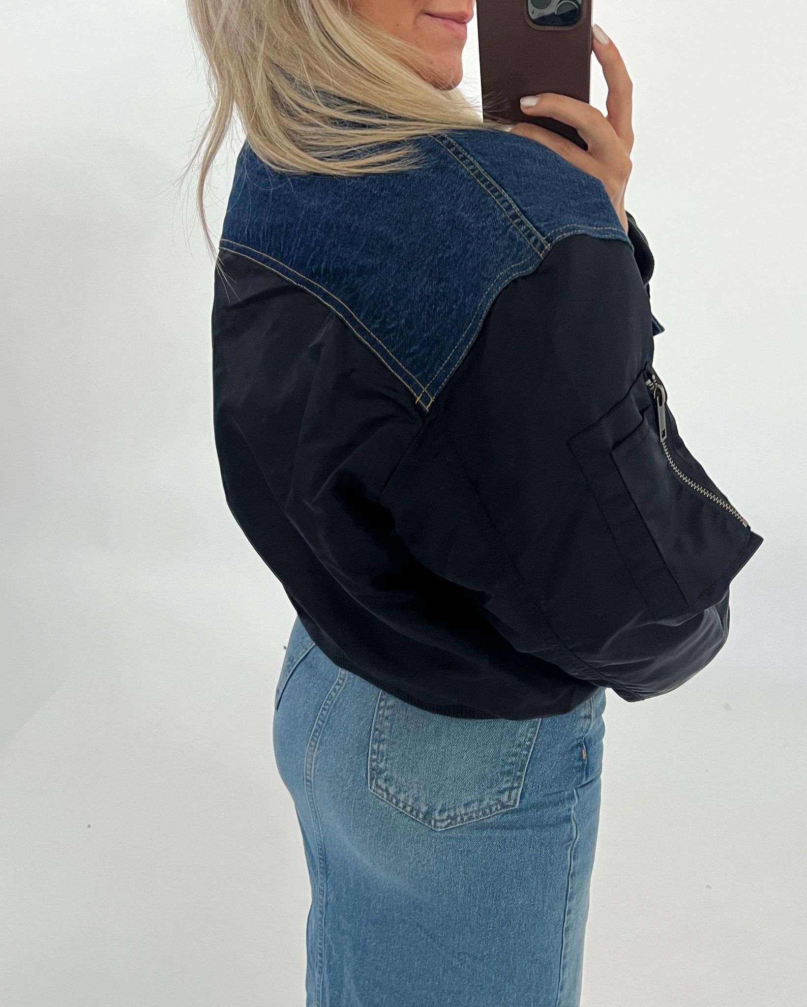 En person står iført en sort og blå jakke og lyst hår, tager et spejlbillede med en smartphone. Personen bærer også en lyseblå denim nederdel. Baggrunden er hvid.