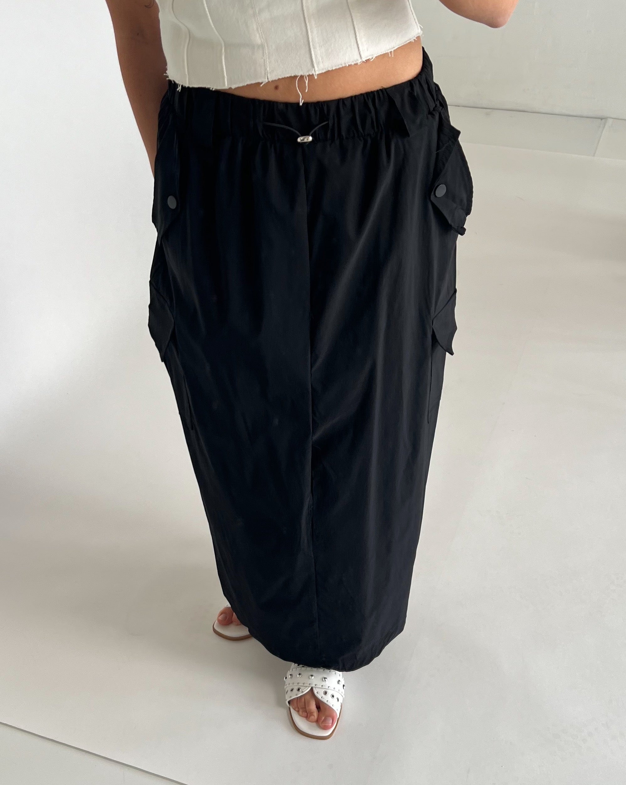 Sort lang nederdel med lommer og elastisk talje bæres af person iført hvid top og sandaler. Personen står på lys baggrund i minimalistisk rum.