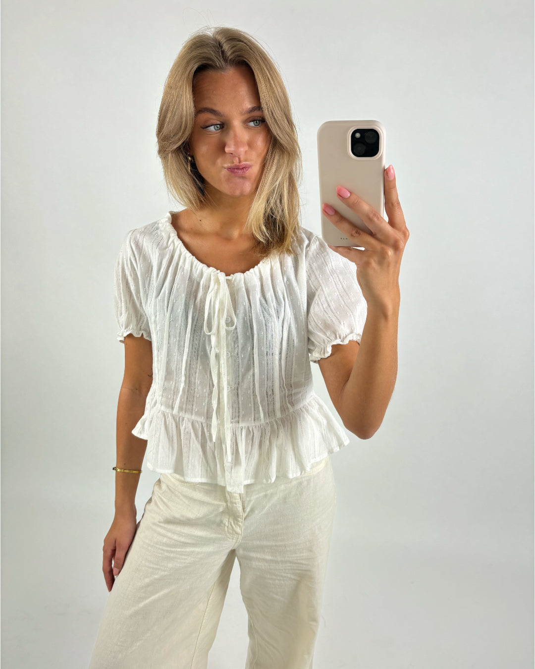 En person tager en selfie med en mobiltelefon iført en hvid top og hvide bukser i et lyst rum uden tekst.