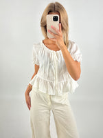 En person tager en selfie foran et spejl. De bærer en hvid bluse og bukser, mens de står i et enkelt, lyst rum. Personen bruger en mobiltelefon med et lyst cover.