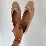 En hånd holder et par beige ballerina-sko med sløjfer, stående mod en hvid baggrund. Inde i skoene står der "stephan PARIS." Hånden har et guldarmbånd og rød snor.