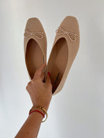 En hånd holder et par beige ballerina-sko med sløjfer, stående mod en hvid baggrund. Inde i skoene står der "stephan PARIS." Hånden har et guldarmbånd og rød snor.