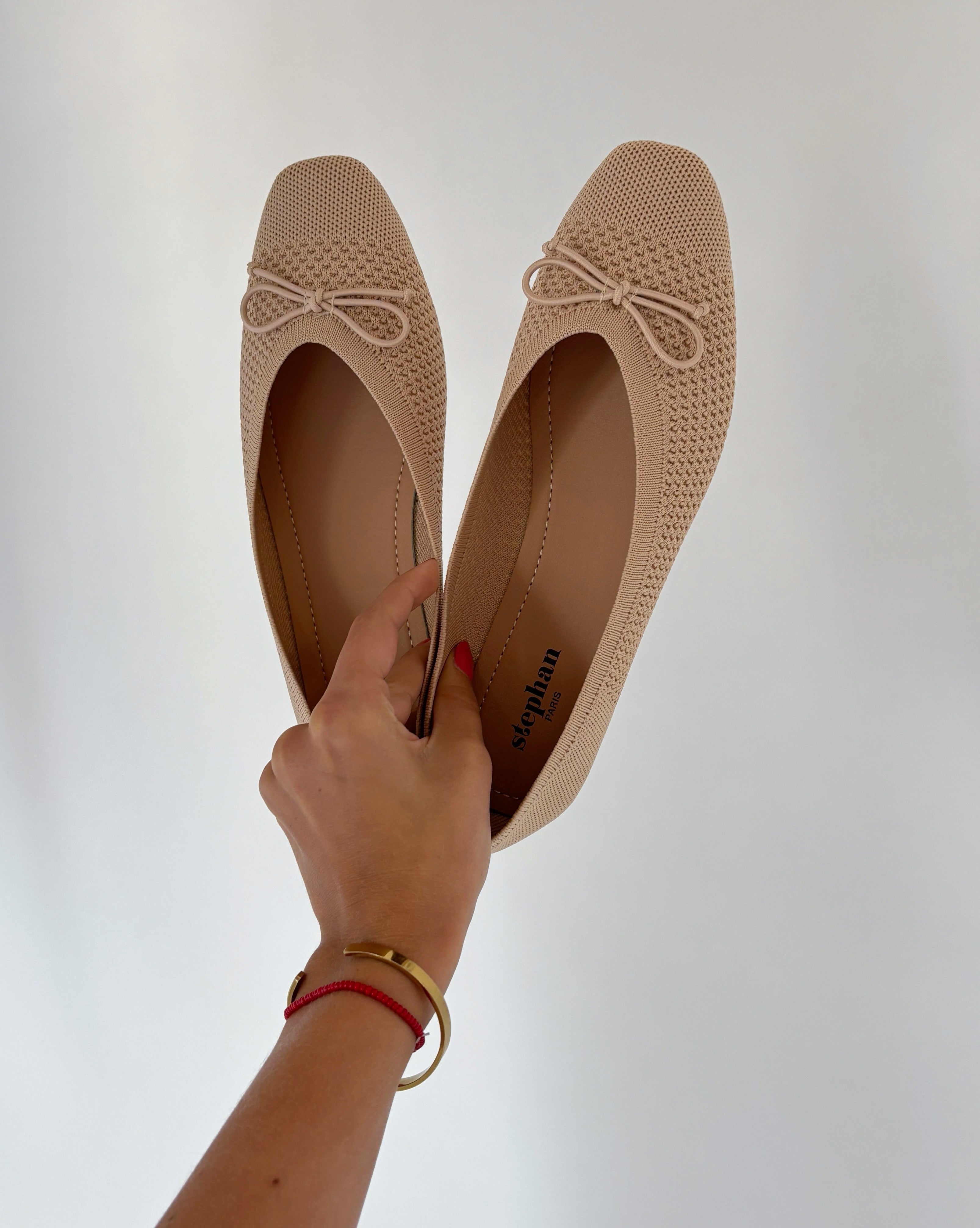 En hånd holder et par beige ballerina-sko med sløjfer, stående mod en hvid baggrund. Inde i skoene står der "stephan PARIS." Hånden har et guldarmbånd og rød snor.