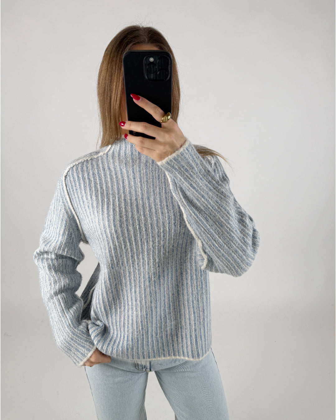 Person iført en stribet sweater og lyse jeans tager et billede med en sort telefon foran sig, i et lyst, neutralt rum.