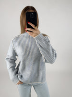 Person iført en stribet sweater og lyse jeans tager et billede med en sort telefon foran sig, i et lyst, neutralt rum.