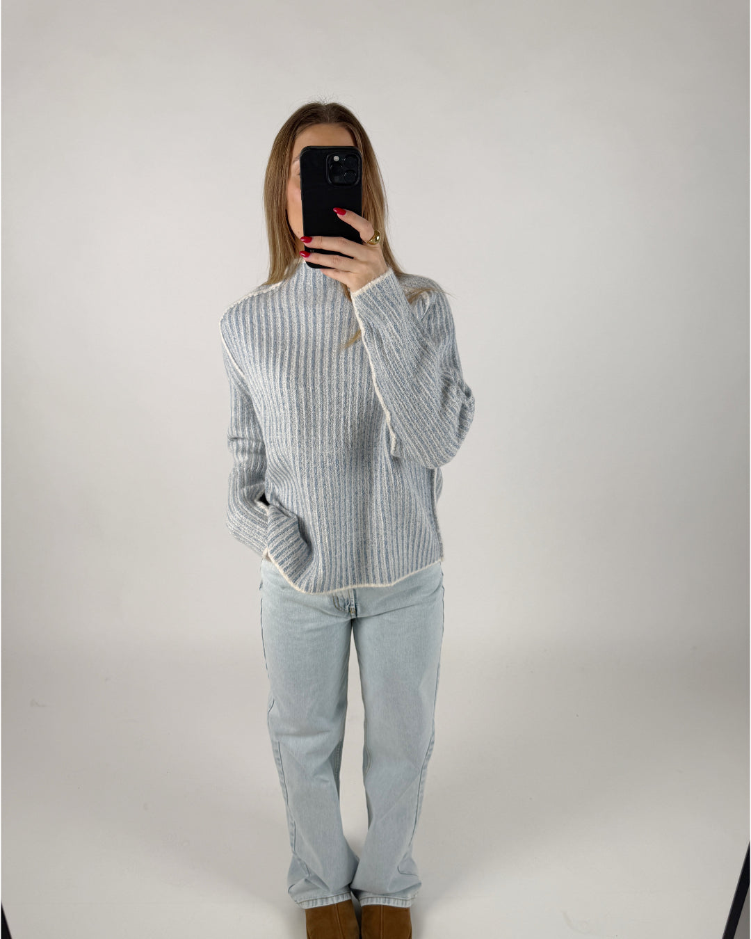 En person iført en lyseblå striktrøje og lyse jeans står i et minimalistisk rum, mens de tager et billede af sig selv med en smartphone.