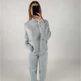 En person iført en lyseblå striktrøje og lyse jeans står i et minimalistisk rum, mens de tager et billede af sig selv med en smartphone.
