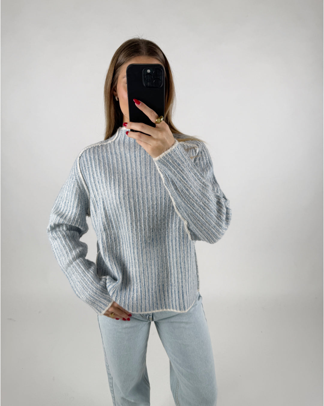 En person i en stribet sweater tager en selfie med en sort telefon. Personen står i et enkelt, hvidt rum og bærer afslappede, lyseblå jeans.