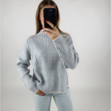 En person i en stribet sweater tager en selfie med en sort telefon. Personen står i et enkelt, hvidt rum og bærer afslappede, lyseblå jeans.