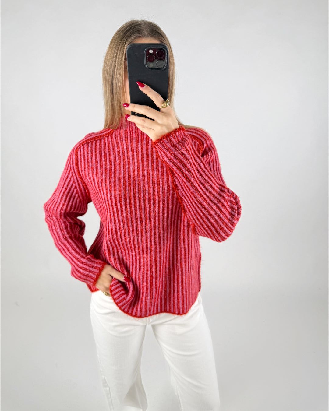 En person iført en rødstribet sweater tager et spejlfoto, mens hånden hviler i lommen på hvide bukser. Baggrunden er enkel og lys.