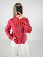 En person iført en rødstribet sweater tager et spejlfoto, mens hånden hviler i lommen på hvide bukser. Baggrunden er enkel og lys.