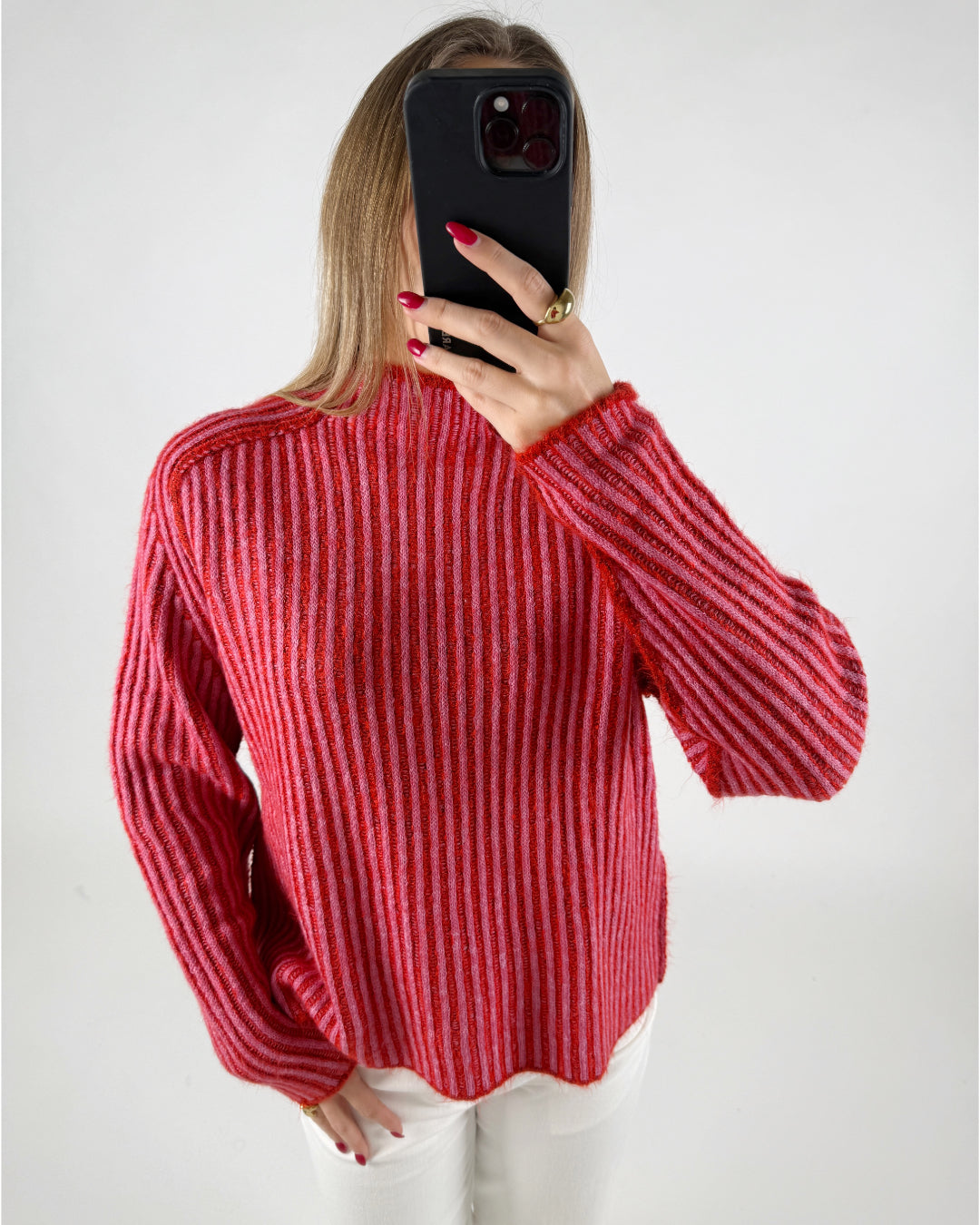 En person iført en rød- og pinkstribet sweater tager et spejlbillede med en sort mobiltelefon. Personen har rød neglelak og bærer en ring. Baggrunden er enkel og lys.
