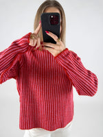 En person holder en sort telefon op foran ansigtet iført en rødstribet sweater, omgivet af en lys baggrund. Personen viser pegefingeren på telefonens skærm.