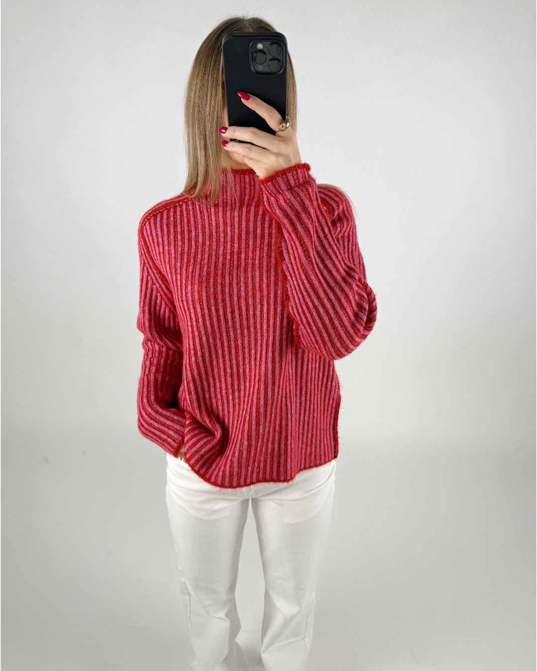 Person fotograferer sig selv, iført en rødstribet sweater og hvide bukser, med en mobiltelefon foran ansigtet. Baggrunden er enkel og lys.