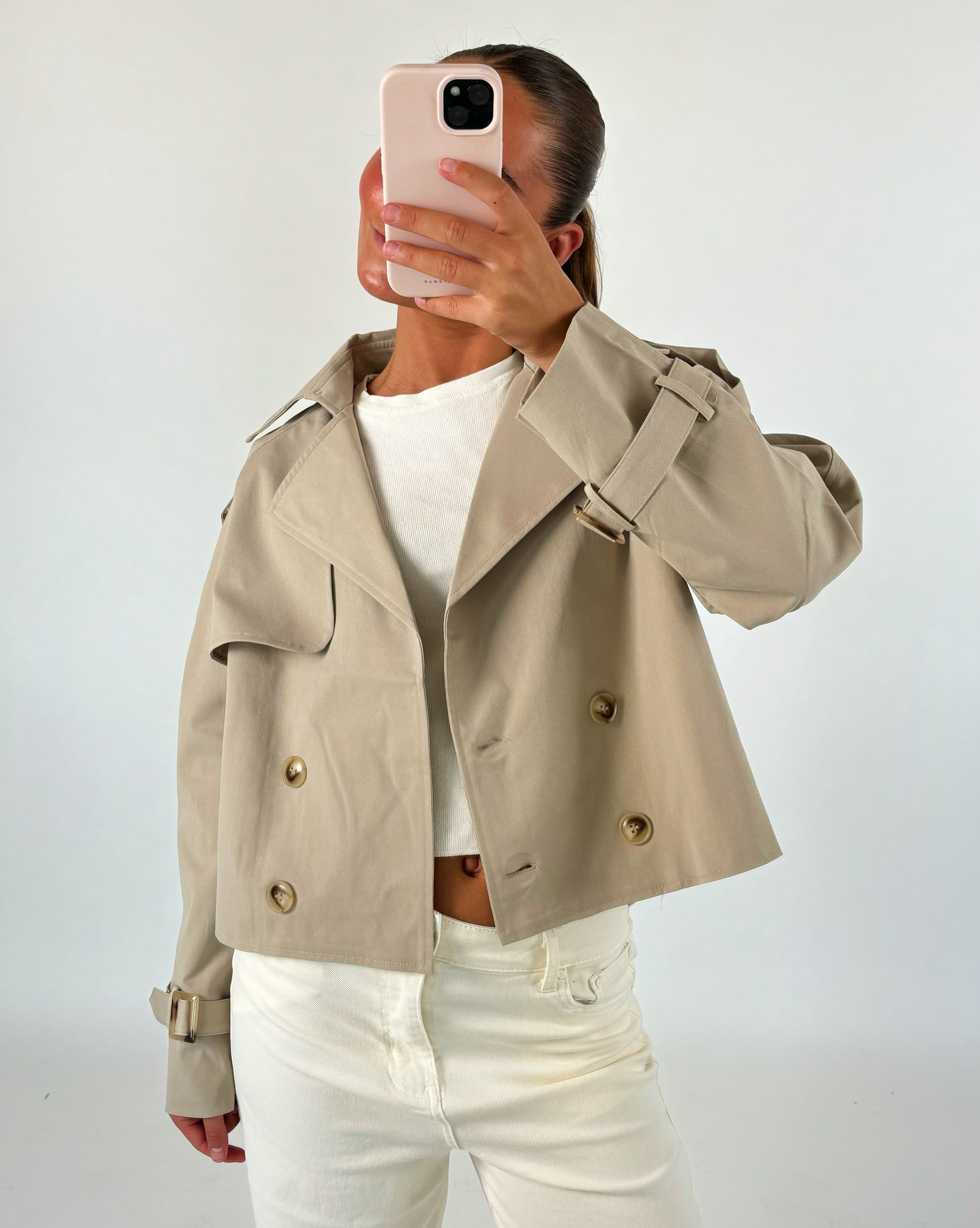 En person i beige jakke og hvide bukser tager et selfie foran en neutral baggrund. Personen holder en lyserød telefon, der dækker ansigtet.