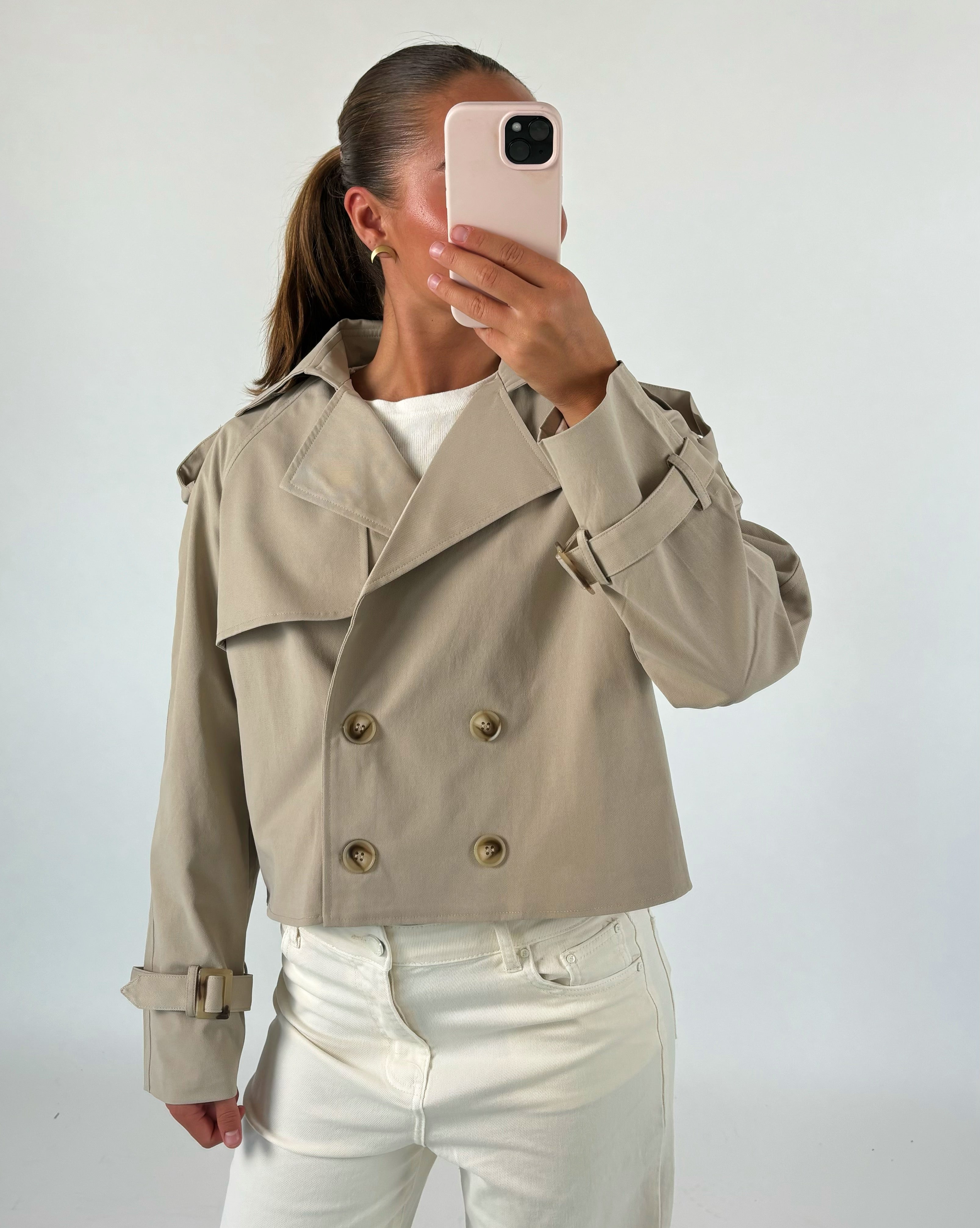 En person tager et spejlbillede iført en beige jakke med knapper og hvid top. De står i et rum med lys baggrund og holder en mobiltelefon foran ansigtet.