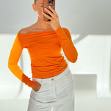 En person iført en orange off-shoulder trøje og hvide bukser tager en selfie i et lyst, hvidt rum. Personen har en telefon i en brun etui.