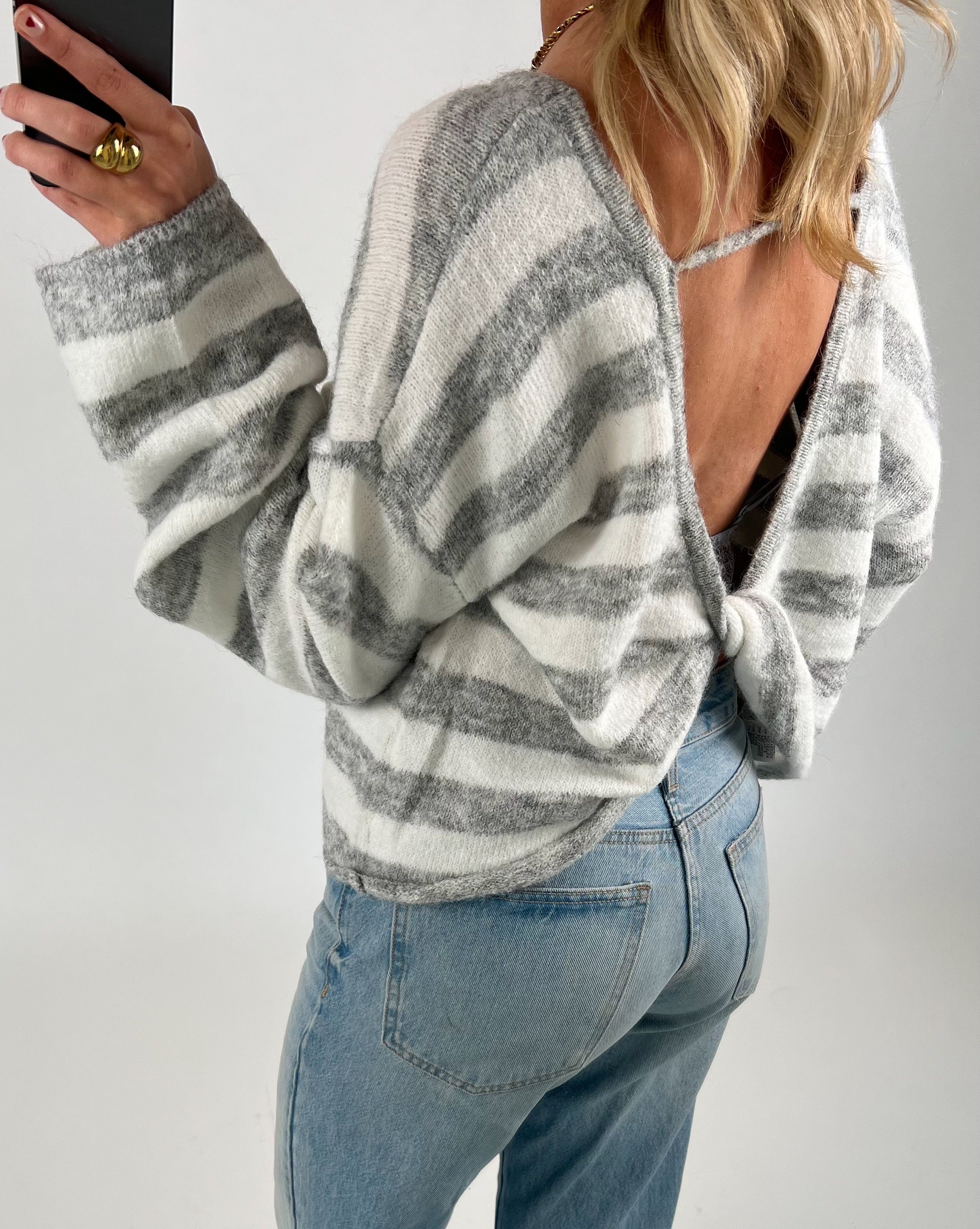 En person iført en stribet hvid og grå trøje med åben ryg samt blå jeans står i en neutral baggrund. Personen holder en sort telefon og viser et guldfarvet smykke på fingeren.