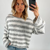 En person tager en selfie iført en stribet grå og hvid trøje og blå jeans. Personen står i et enkelt, lyst rum og kigger til siden.