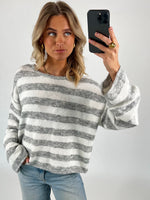 En person tager en selfie iført en stribet grå og hvid trøje og blå jeans. Personen står i et enkelt, lyst rum og kigger til siden.