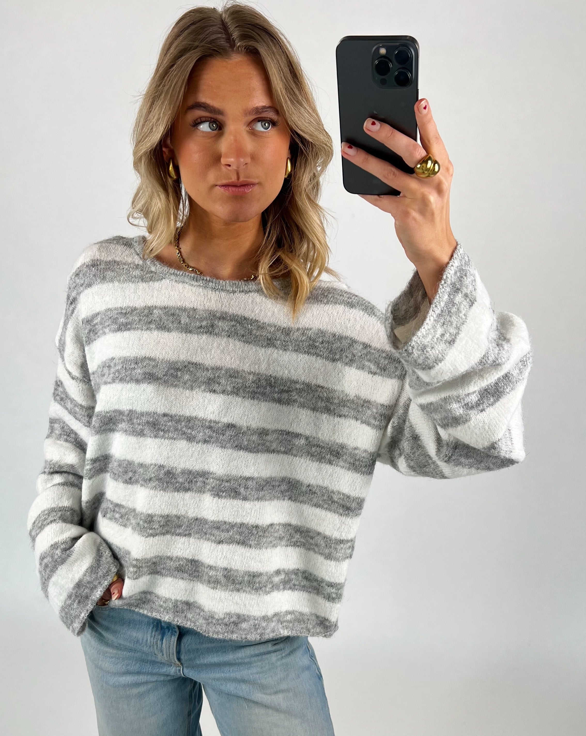 En person tager en selfie iført en stribet grå og hvid trøje og blå jeans. Personen står i et enkelt, lyst rum og kigger til siden.