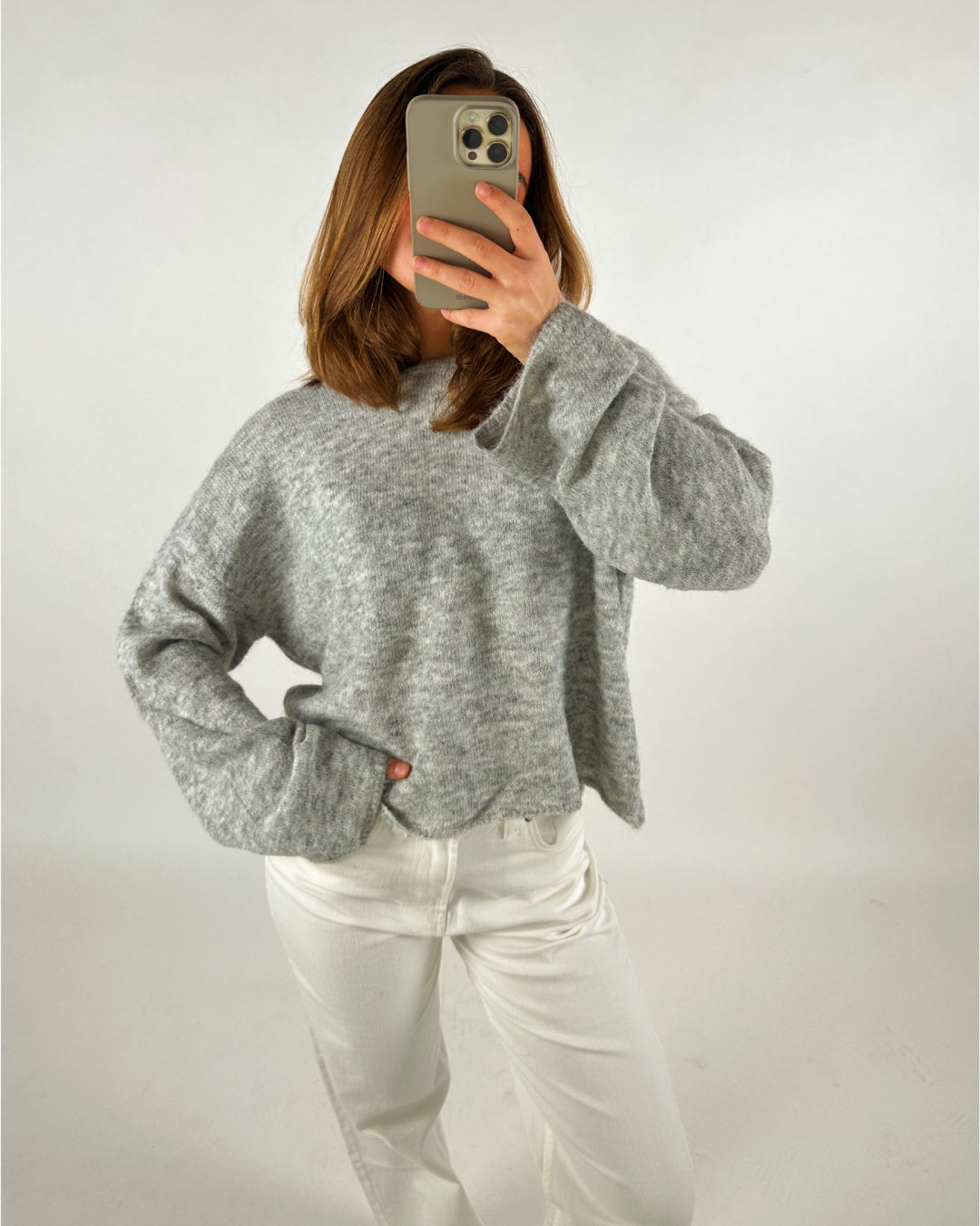 En person iført en grå sweater tager et spejlbillede med en smartphone i hånden, omgivet af en ensfarvet lys baggrund.