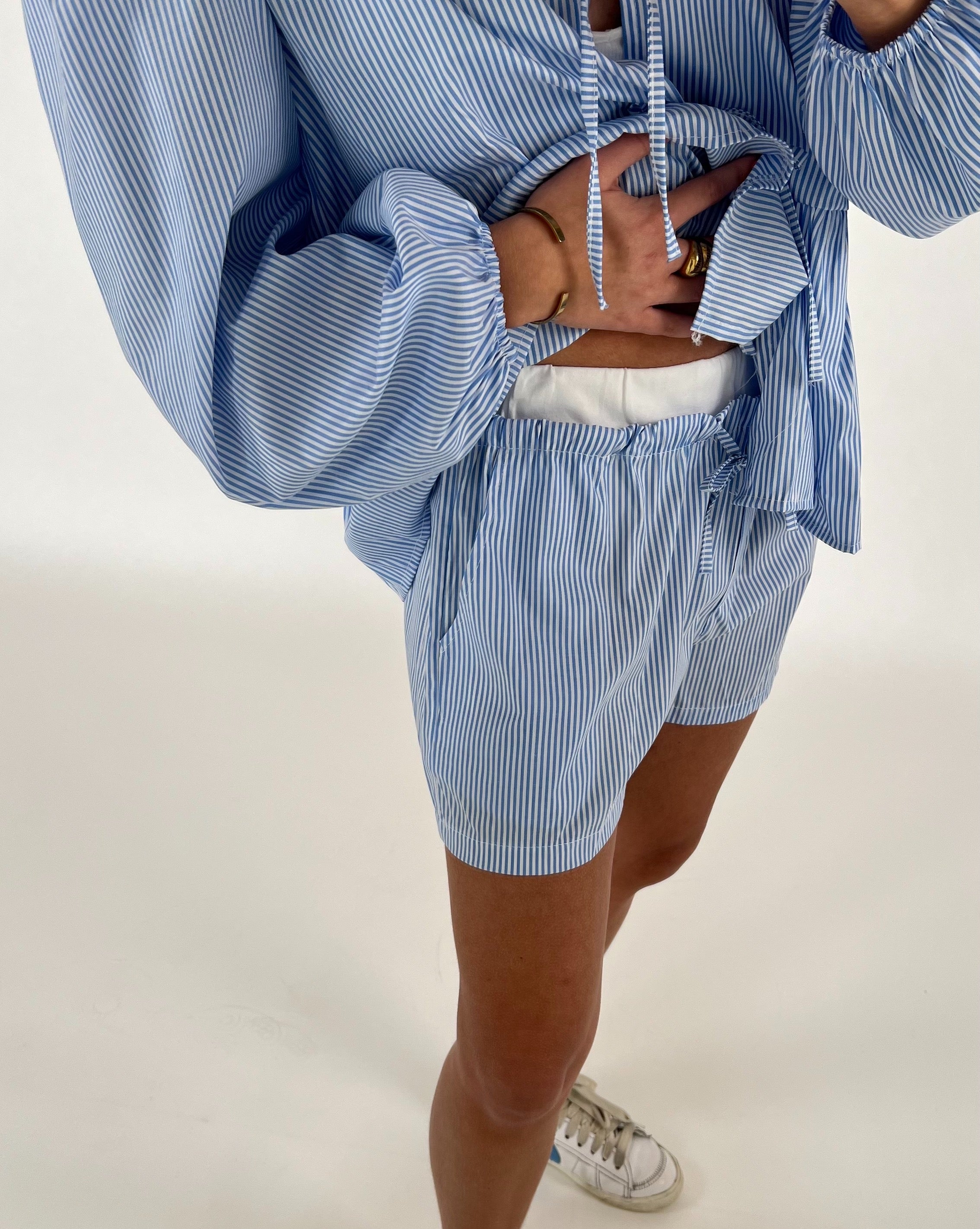 En person iført lyseblå og hvidstribet sæt, bestående af en skjorte og shorts, står med afslappet kropsholdning. Personen er indendørs, iført hvide sneakers.