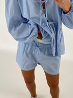Person iført blå- og hvidstribet sæt med shorts og langærmet top, knyttet i taljen. Personen står indendørs i et hvidt rum.