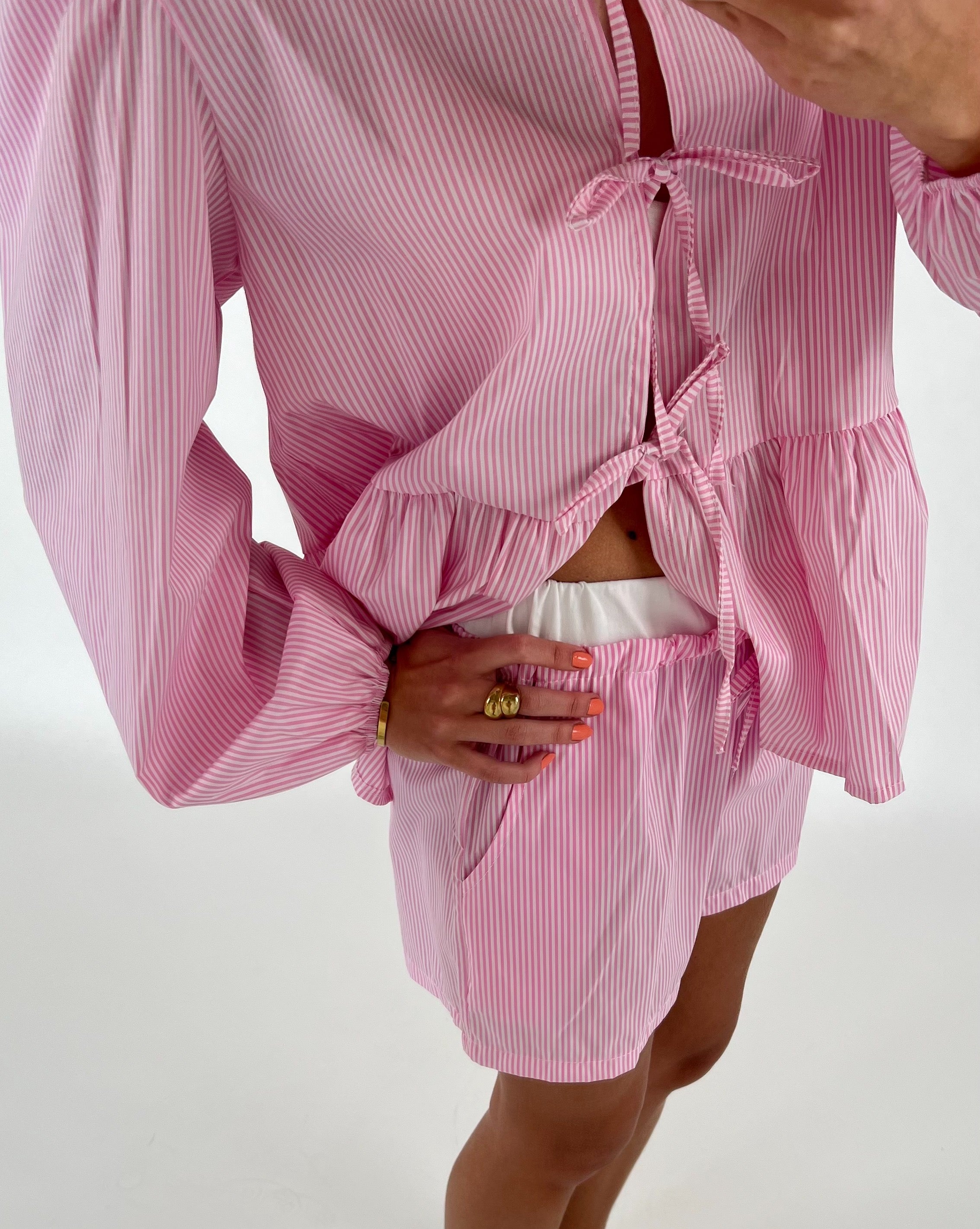 Pink- og hvidstribet sæt med langærmet skjorte og shorts, en person poserer med hånd i lommen. Indendørs hvid baggrund.
