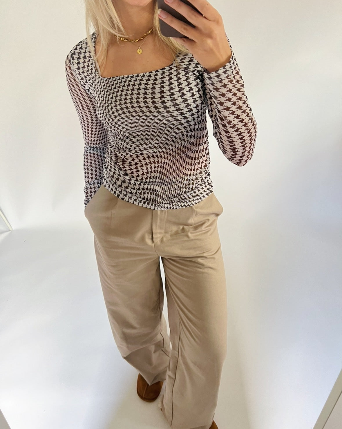 En person iført houndstooth-mønstret top og beige bukser står og tager et spejlbillede med en smartphone i et lyst rum.