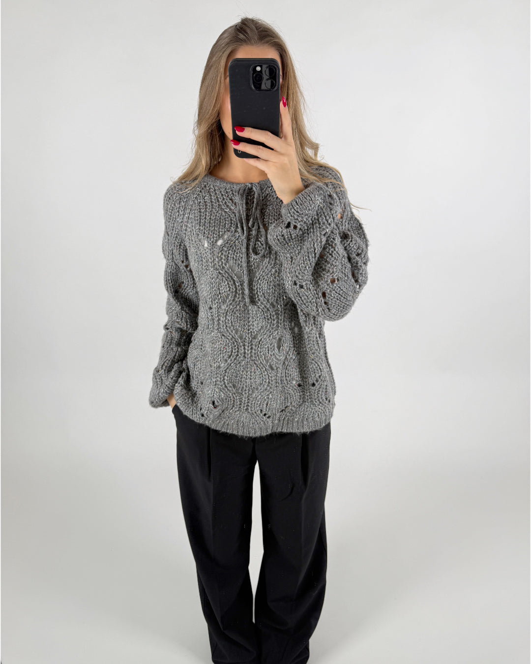 En person iført en grå, strikket sweater holder en sort telefon foran ansigtet. Personen står i et neutralt rum med en hvid baggrund.