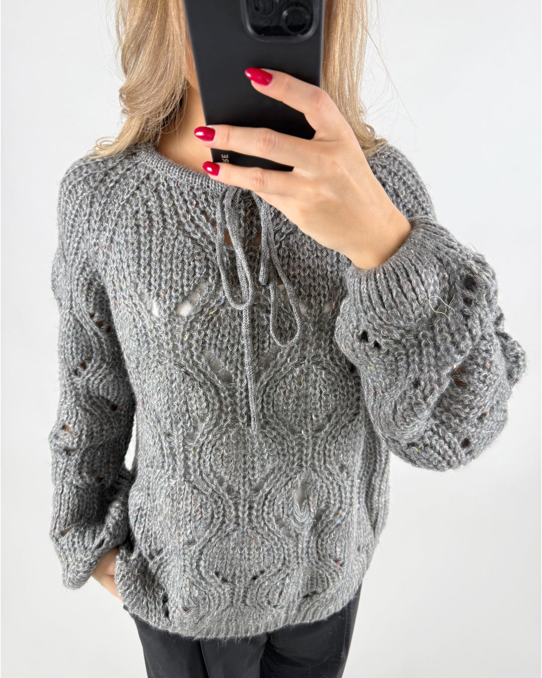 En person iført en grå, strikket sweater tager en selfie med en sort smartphone. Sweateren har snørebånd og et åbent mønster. Personen har røde negle, og baggrunden er neutral.