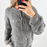 En person iført en grå, strikket sweater tager en selfie med en sort smartphone. Sweateren har snørebånd og et åbent mønster. Personen har røde negle, og baggrunden er neutral.